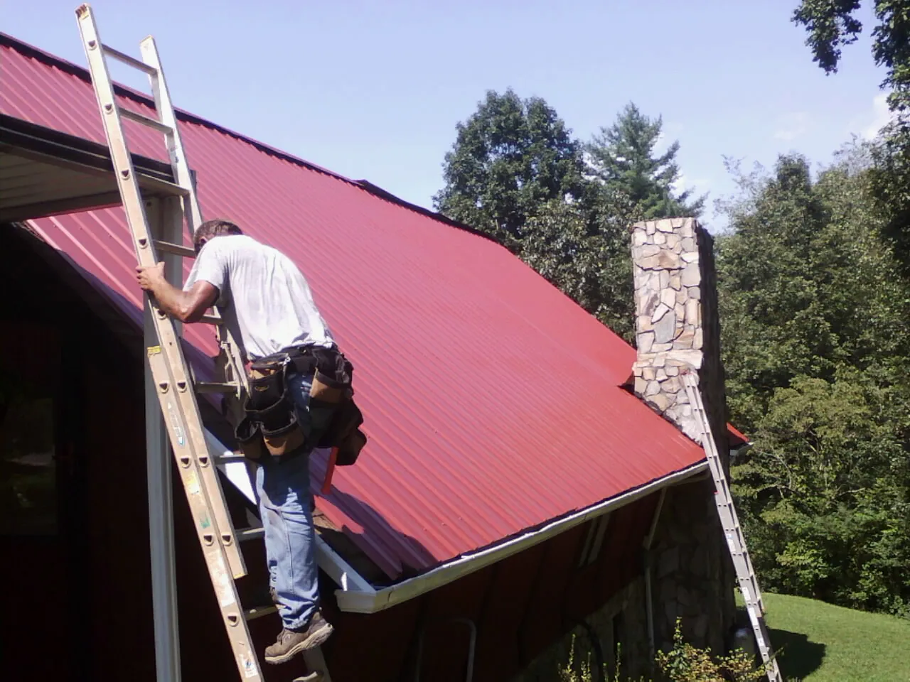 Roofer on a ladder beside a red metal roof and stone chimney — Soffit & Fascia Repair by Roofing Brothers in Coatesville